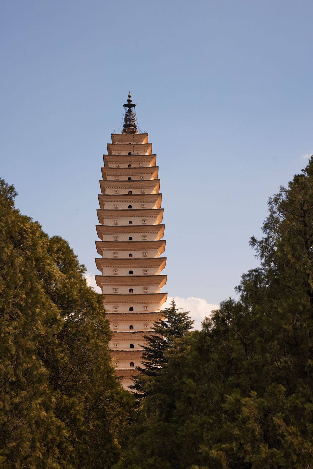 Ancient pagoda tower with tiered architecture rises above trees against blue sky in traditional Chinese style.