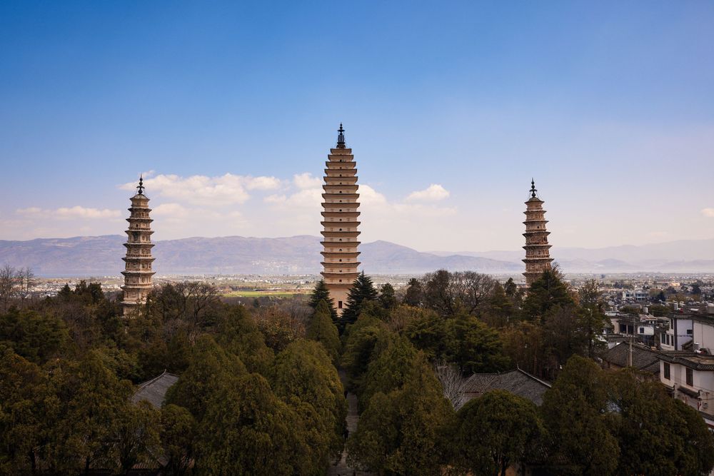 Three ancient Buddhist pagoda towers rise above a forested area in Dali, China against a blue mountain backdrop.