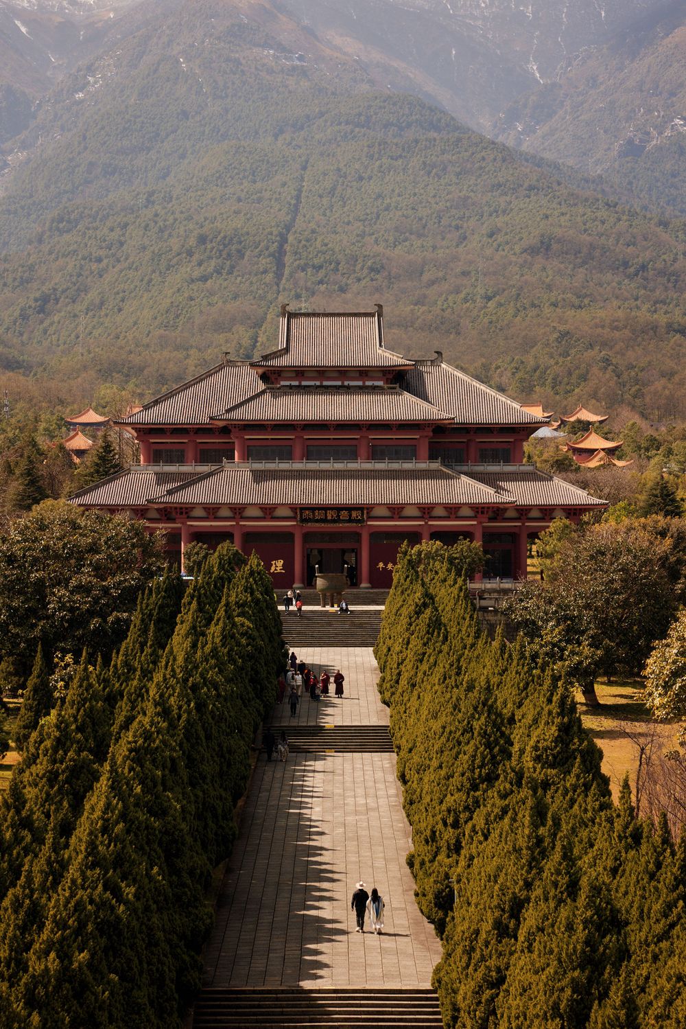 Scenic walkway leading to a traditional Chinese Buddhist temple surrounded by evergreen trees and mountains in the background.