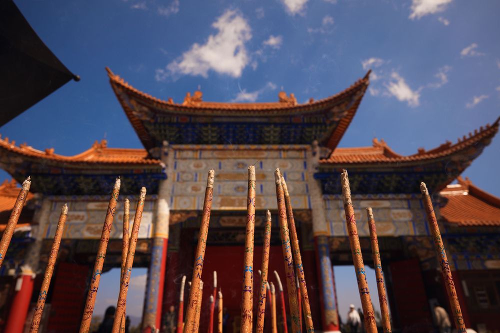 Traditional Chinese temple building with upturned eaves and tall incense sticks against a blue sky with white clouds.