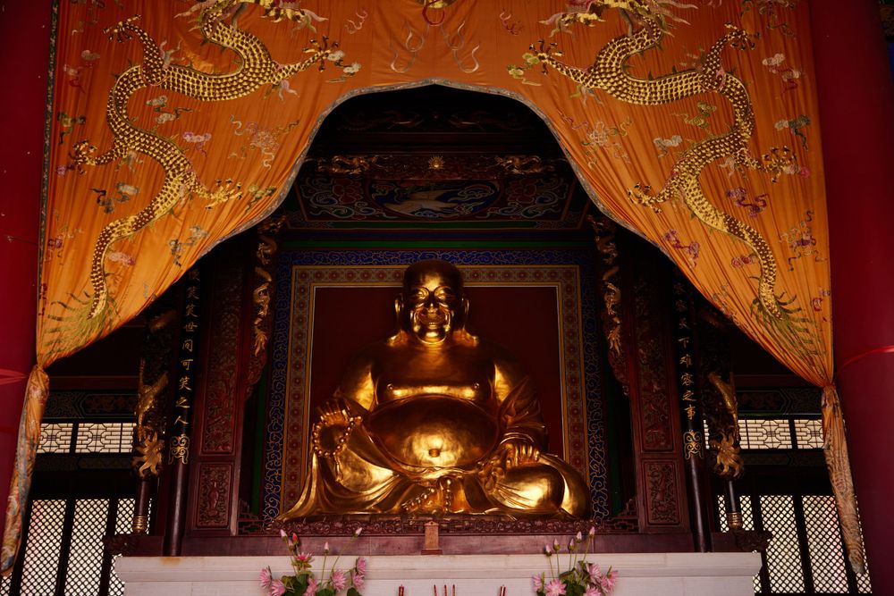 Golden Buddha statue sits in ornate temple shrine with red walls and decorative gold curtains.