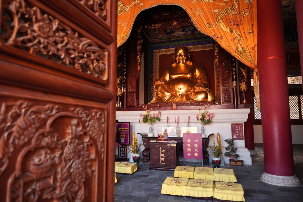 Interior view of a Buddhist temple showing a golden Buddha statue on an altar with ornate wooden carvings and yellow cushions.