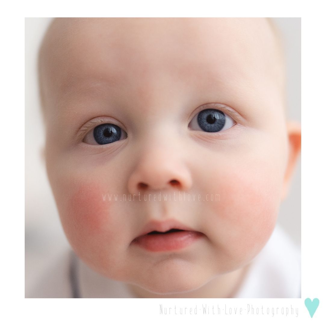 Close-up portrait of a baby with bright blue eyes, rosy cheeks, and soft features against a light background.