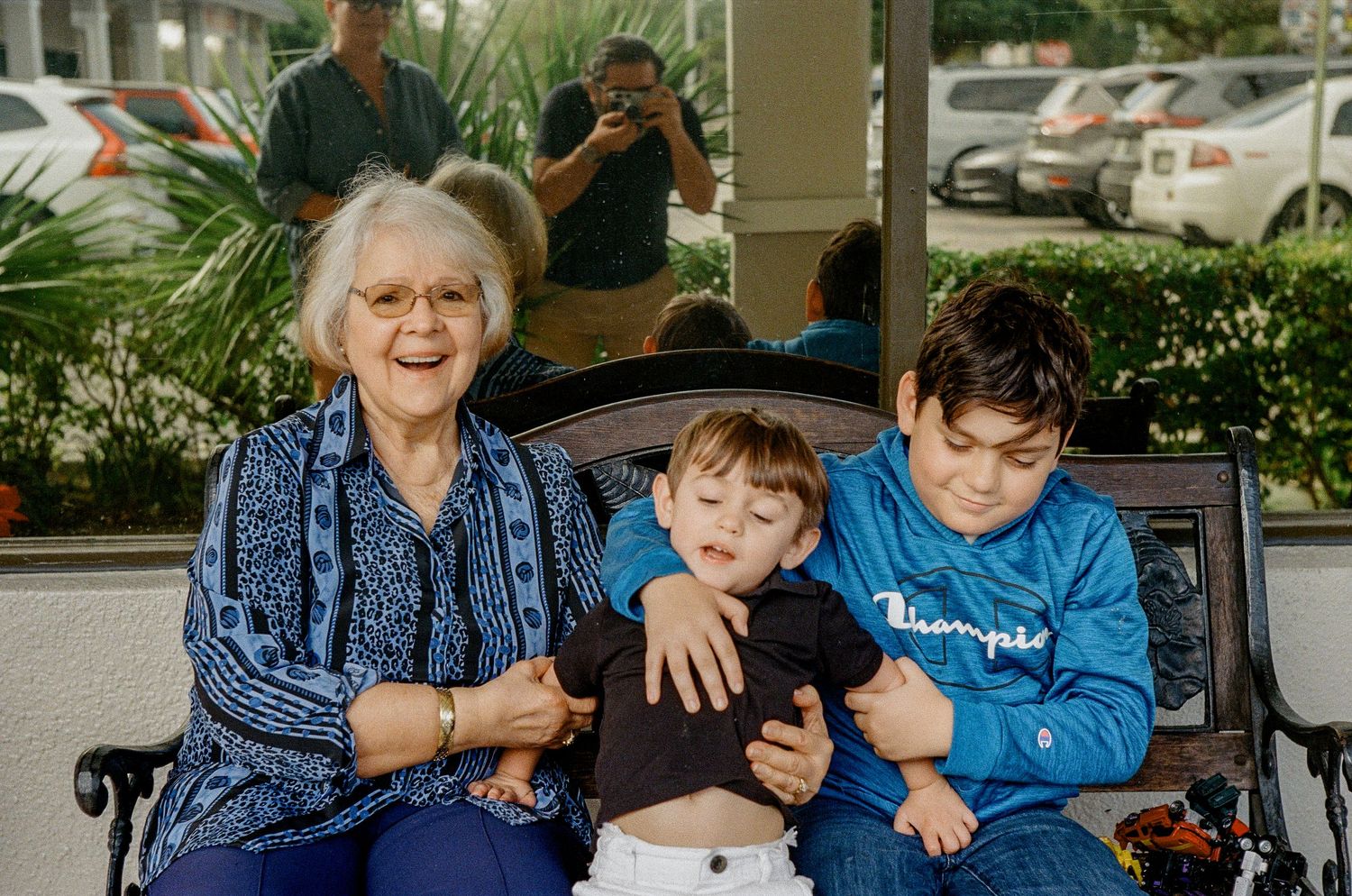 A family sits together on a bench outside, sharing a casual moment at what appears to be a shopping center.