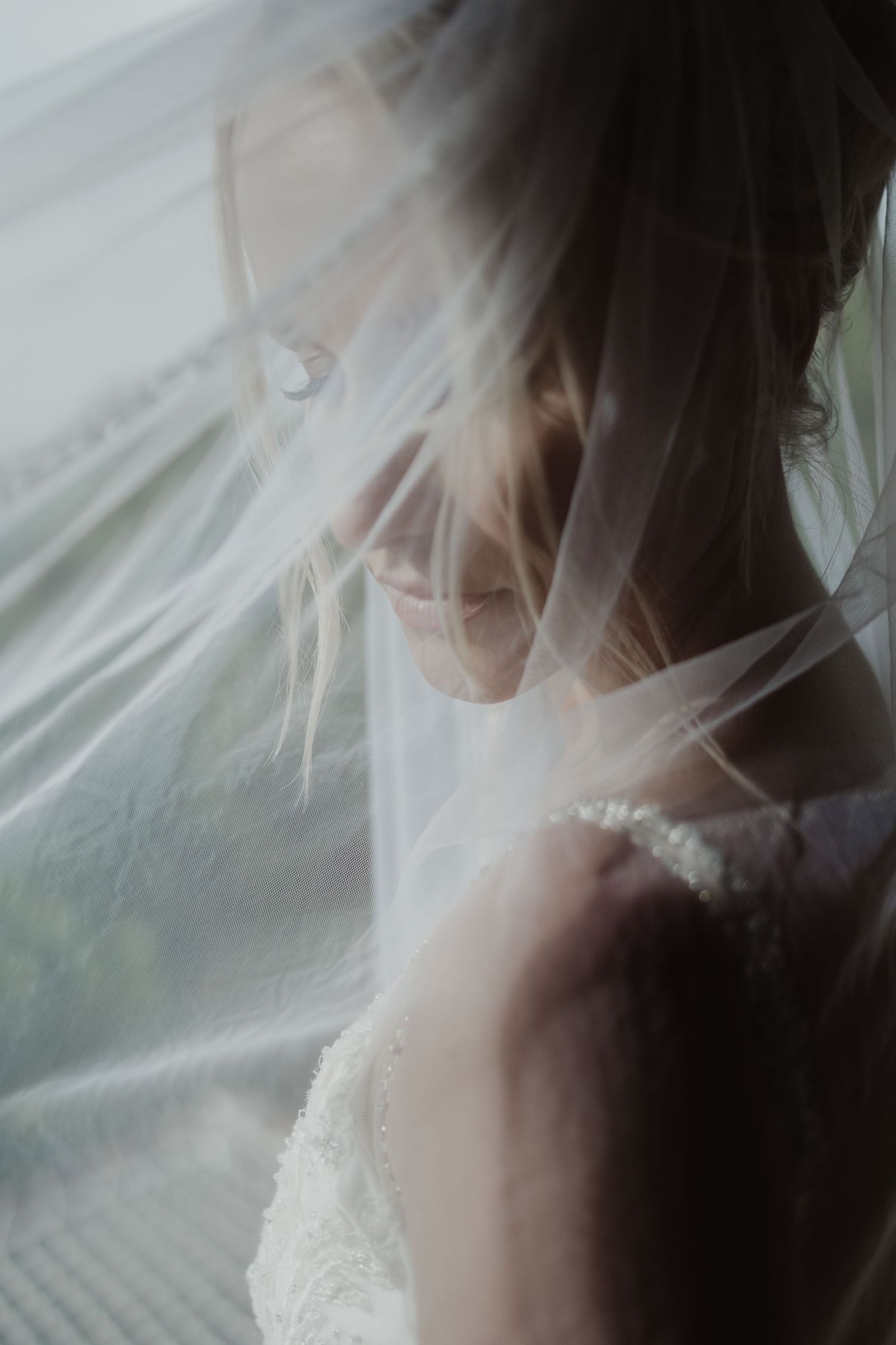 A bride draped in a flowing sheer veil, softly lit near a window in an ethereal wedding portrait.