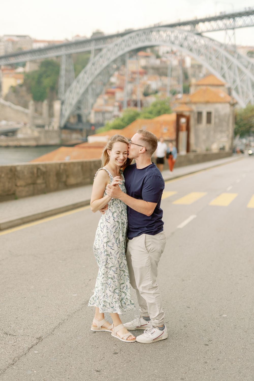 Couple embracing on street with iconic Porto bridge in background.