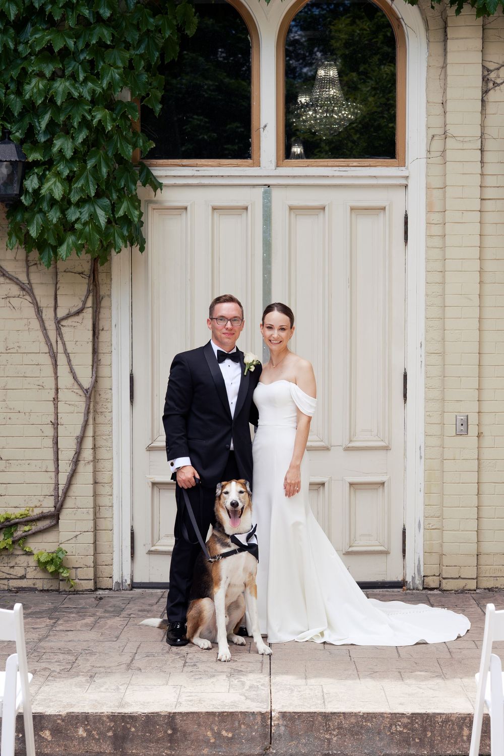A couple in formal wedding attire poses with their black and white dog in front of an elegant cream-colored doorway.