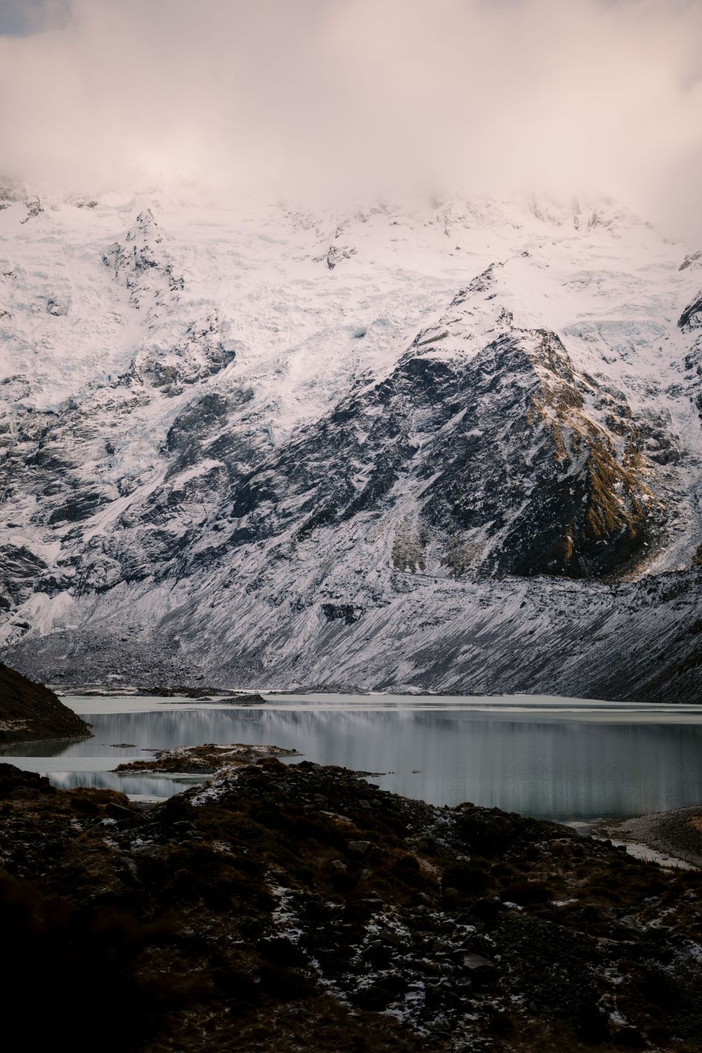 Snow-capped mountains reflected in a serene alpine lake with moody overcast skies.