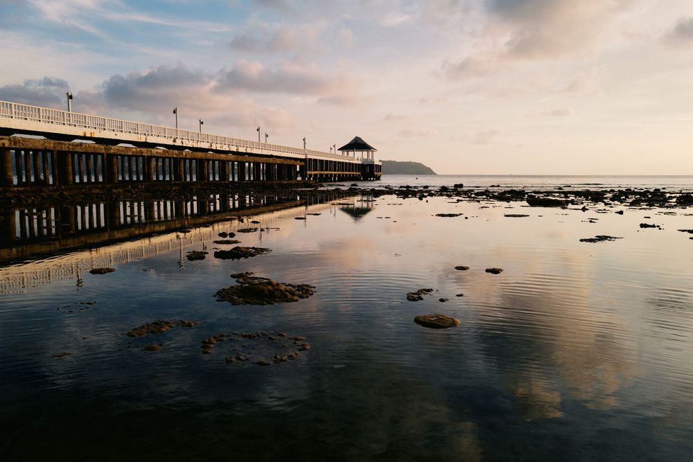 A wooden pier extends over calm tidal waters with rocks and reflections during sunset at the beach.