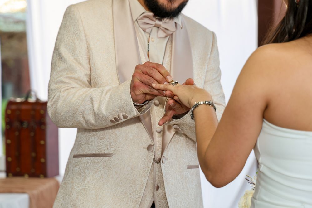 A person in a beige suit and white shirt holds a glass while standing at a formal event.