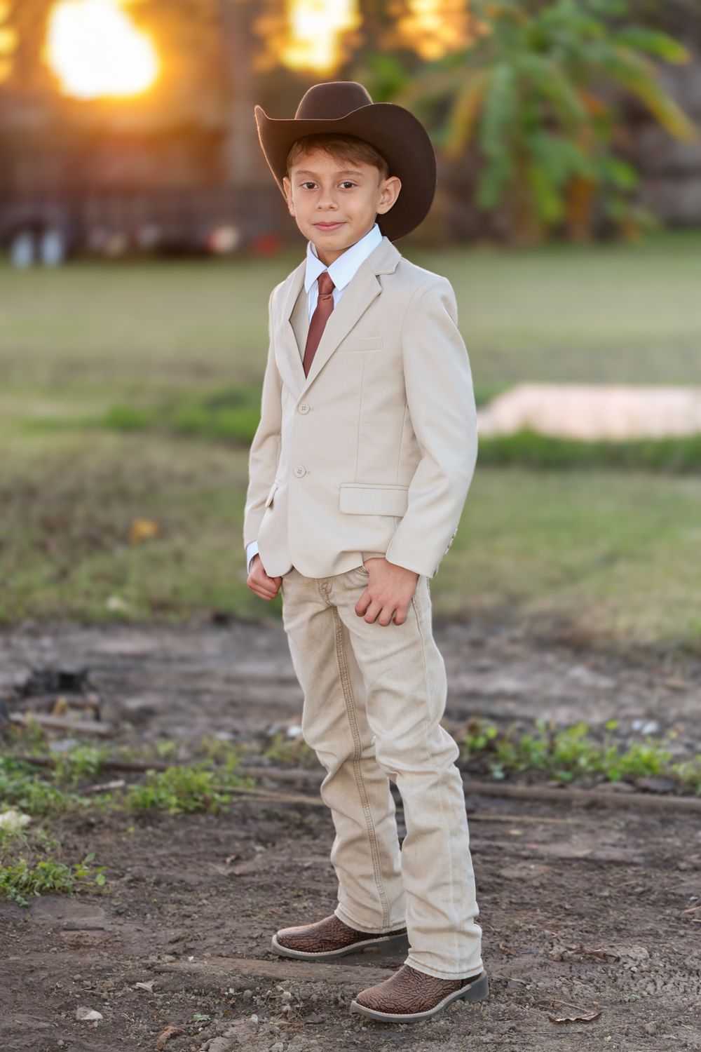 A young figure in a beige suit and brown cowboy boots stands outdoors during golden hour lighting.