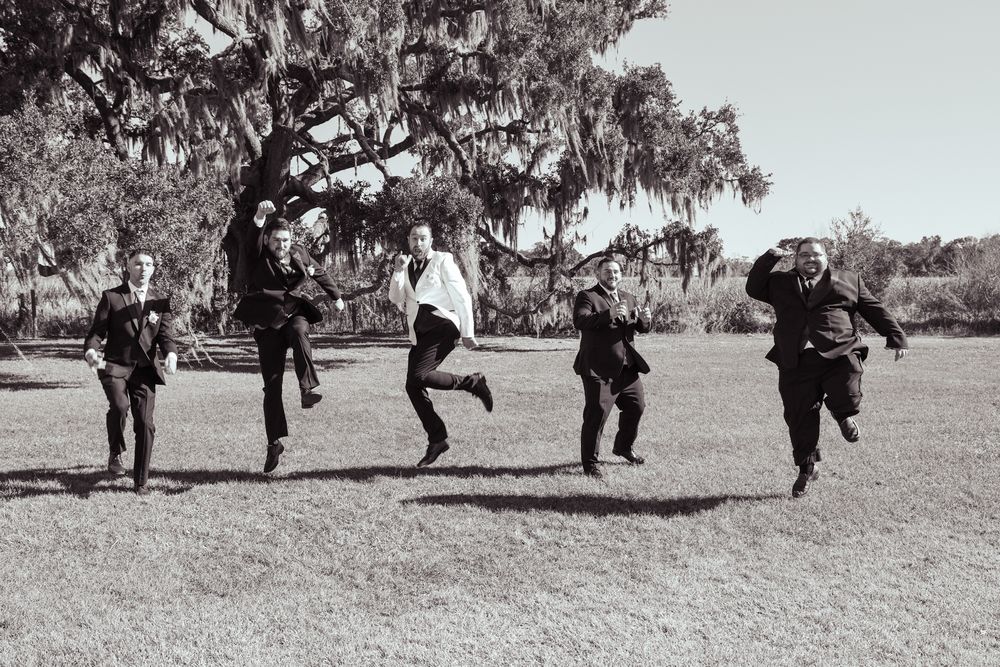 Five groomsmen joyfully jumping in a grassy field beneath a large oak tree in a black and white wedding photo.