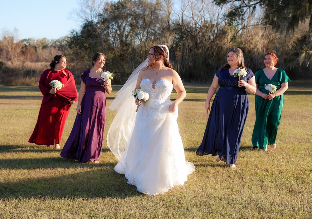 Bride in white gown walks with four bridesmaids in colorful dresses on green lawn at outdoor wedding ceremony.