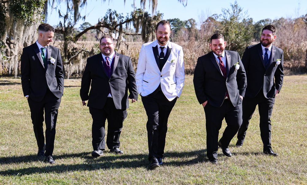 Five groomsmen in black suits walk outdoors on grass, with the groom in a white jacket at center.
