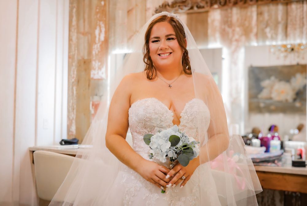 Smiling bride in strapless lace wedding gown holding white bouquet with veil, standing in bridal suite.