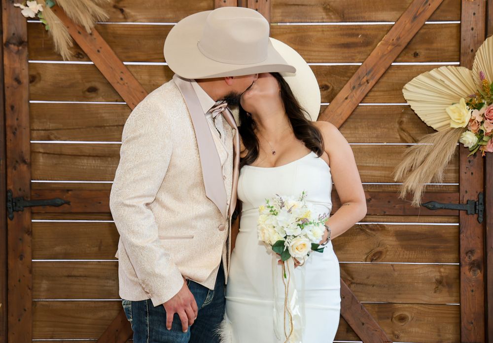A romantic wedding couple in white attire share an intimate moment against a rustic wooden wall backdrop.