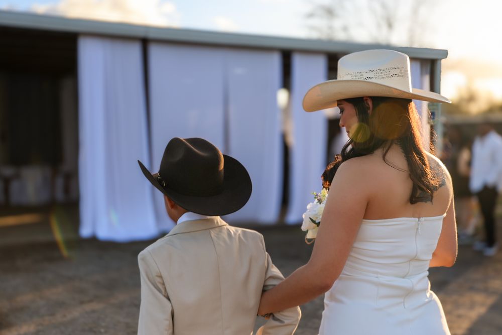 A bride in a white dress and cowboy hat stands with a young person wearing a black cowboy hat at sunset.