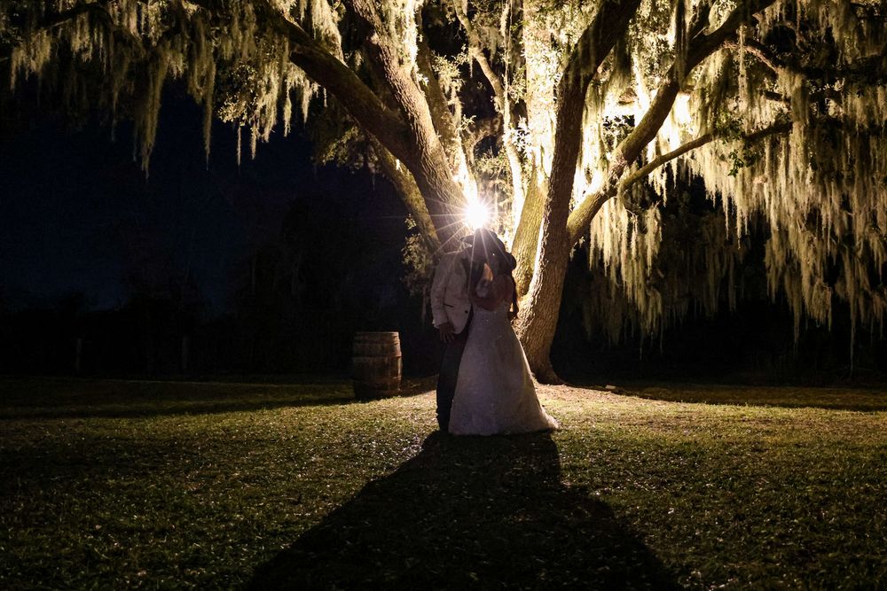 A bride and groom share a romantic moment under a moss-draped oak tree illuminated by a glowing light at night.