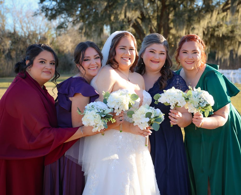 Bride in white gown poses with four bridesmaids in colorful dresses holding white bouquets outdoors.