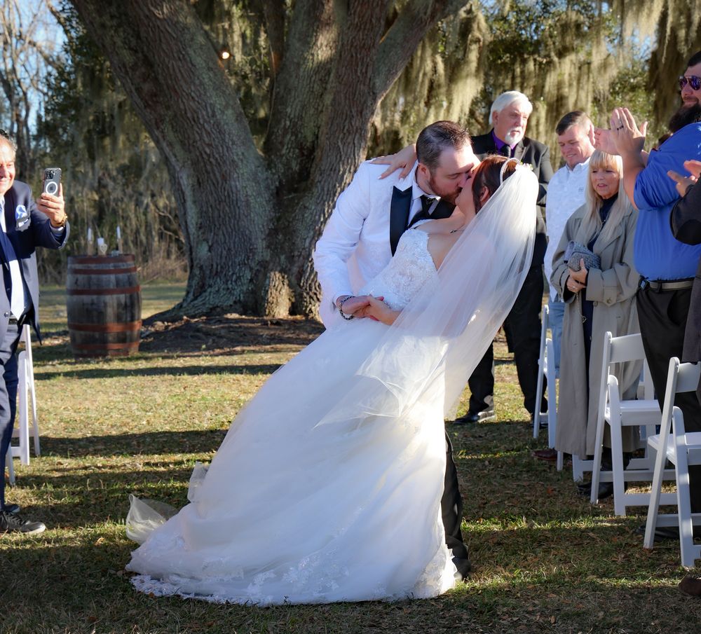 Groom dips bride for a kiss at outdoor wedding ceremony while guests applaud under a large oak tree.