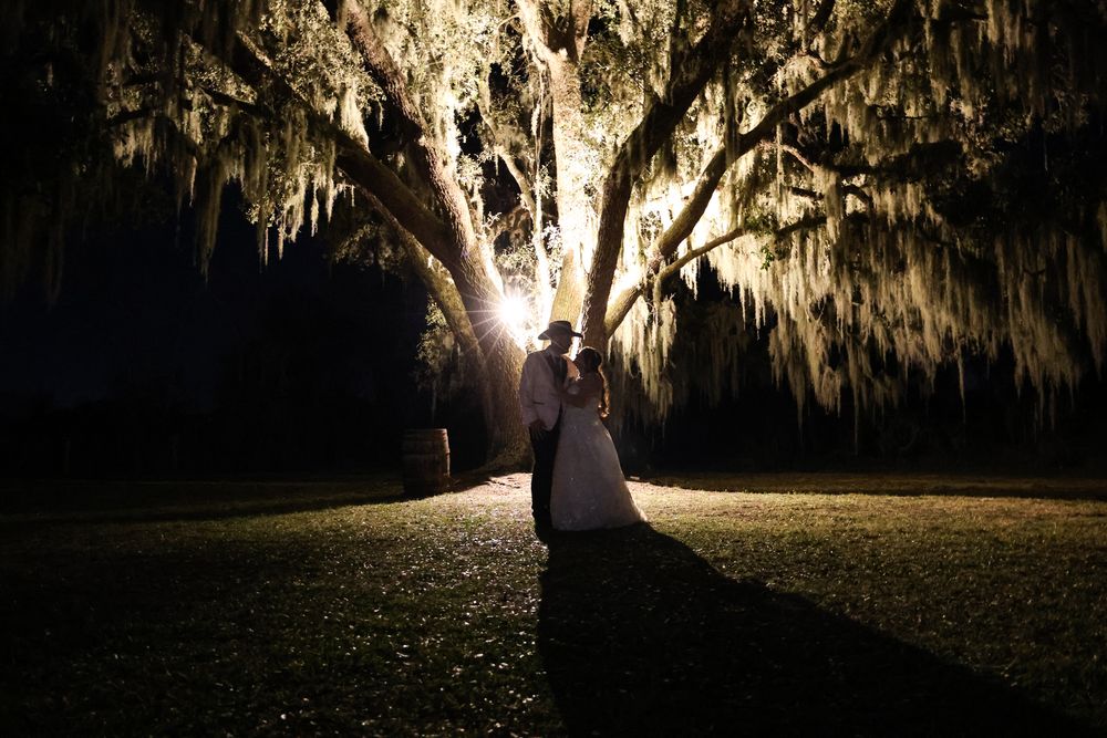Bride and groom sharing romantic moment under a large moss-draped oak tree at night.