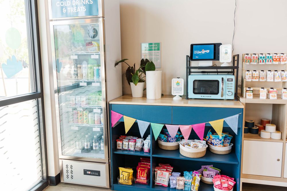 Blue snack station with pennant banner, drink refrigerator, microwave, and colorful packaged treats for kids.