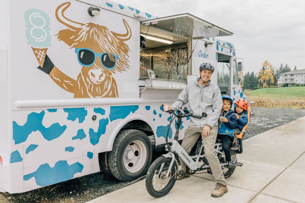 A man and child on bikes pose beside the Eight Cow Creamery food truck with highland cow branding.