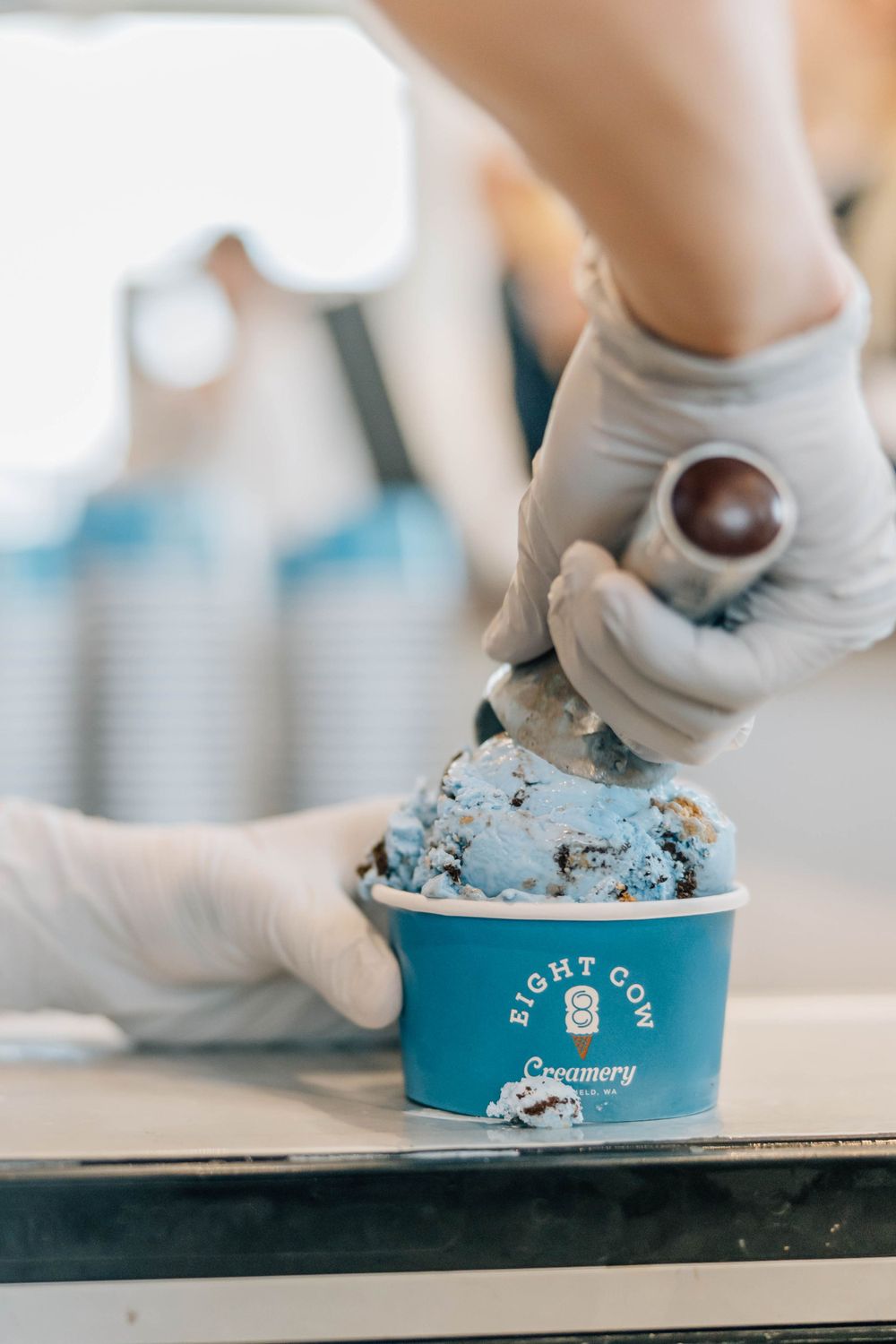 A gloved employee scoops blue ice cream into an Eight Cow Creamery branded cup with a chocolate ball.
