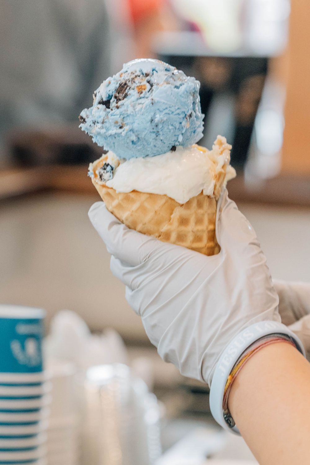 Gloved hand holding a waffle cone with two scoops of blue and white ice cream at an ice cream shop.