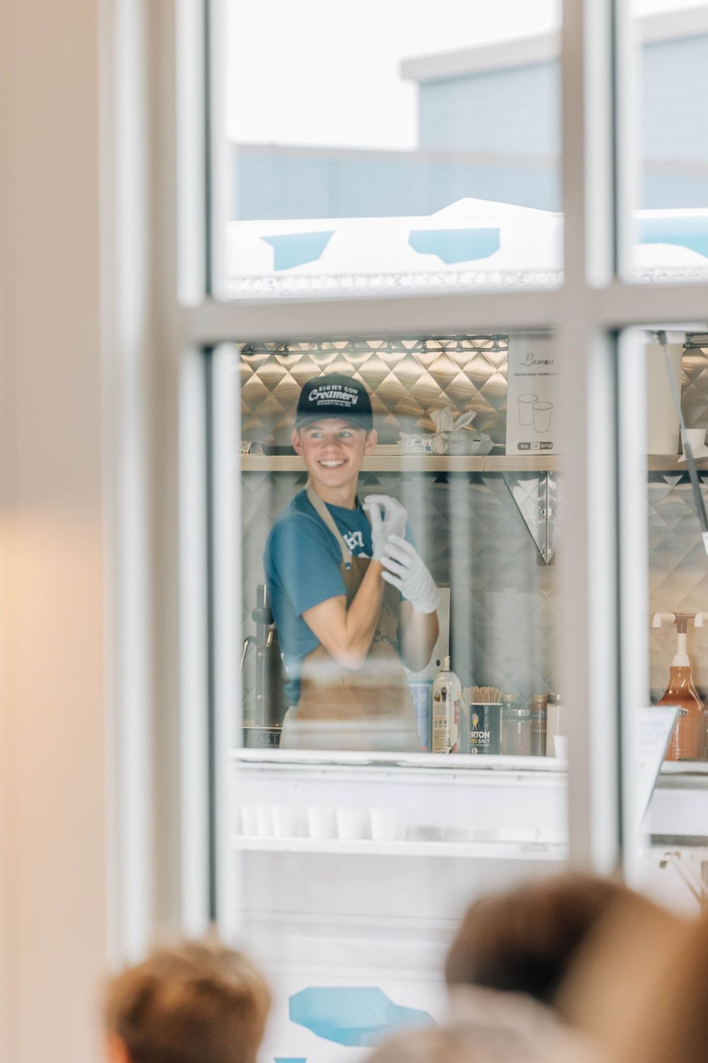 Smiling ice cream shop employee in cap and gloves seen through a window at Eight Cow Creamery.