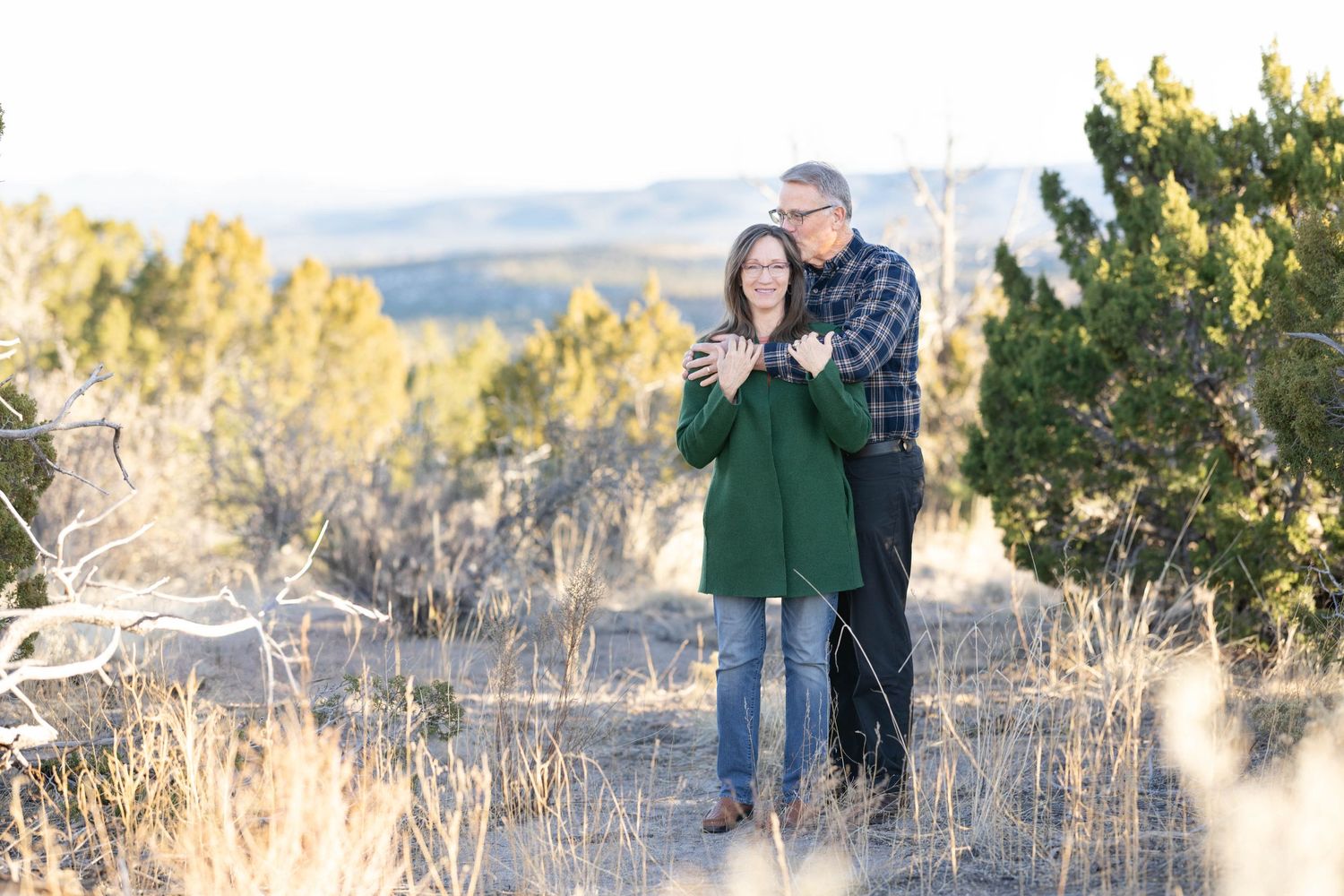 A couple embraces while standing in a field of dry grass with mountains in the distance.
