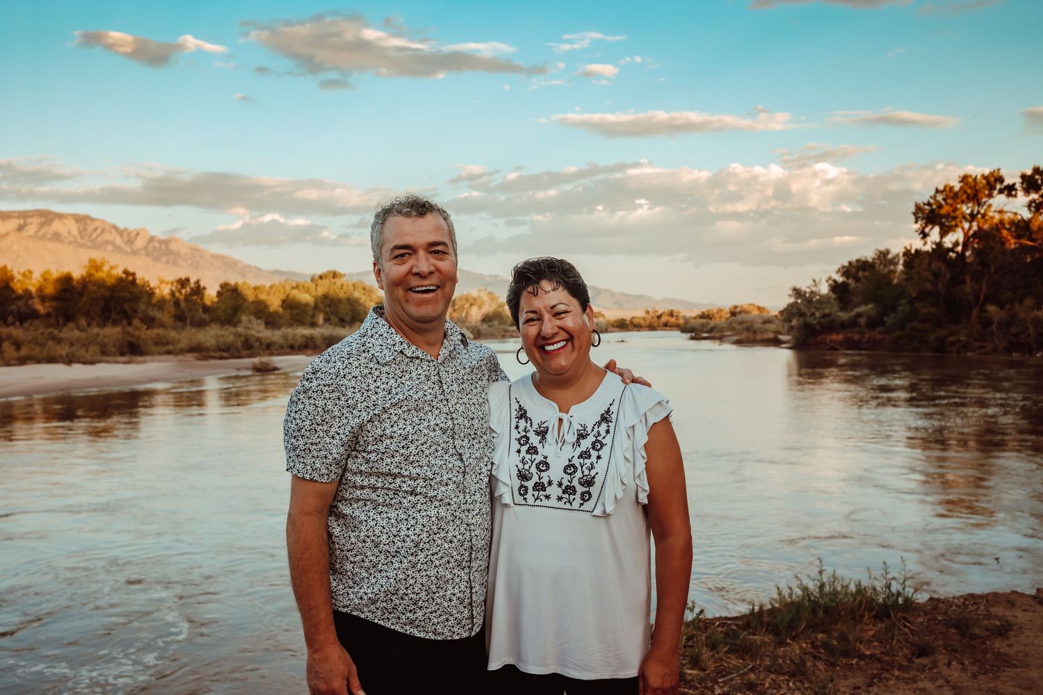 Couple poses together at sunset by scenic river with mountains in background.