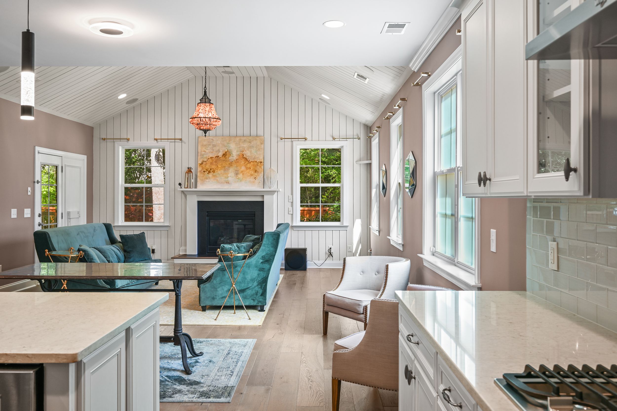 Modern open concept living space with vaulted ceiling features teal accent chairs and white shiplap walls near bright kitchen.