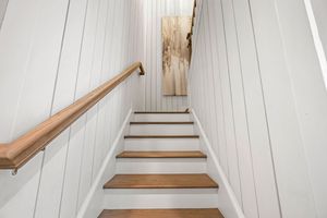 Modern white staircase with wooden treads and handrail leads upwards between vertical shiplap paneled walls.