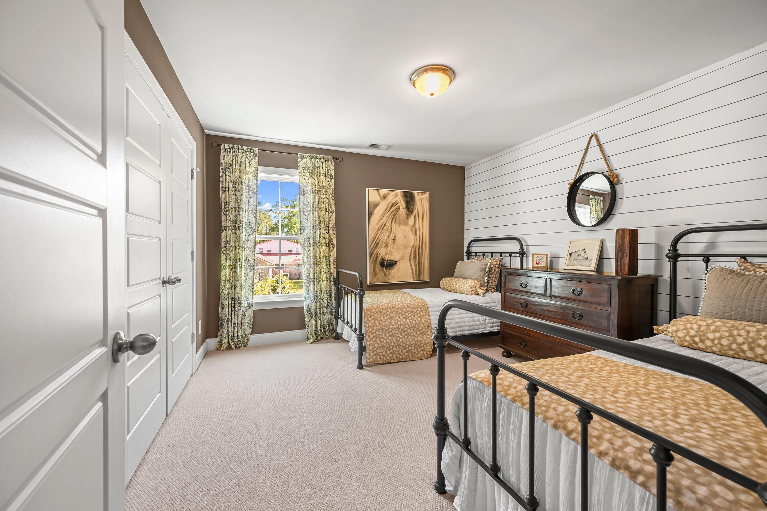 Light-filled bedroom with twin metal beds, shiplap accent wall, floral curtains, and mirror shows mountain views through window.