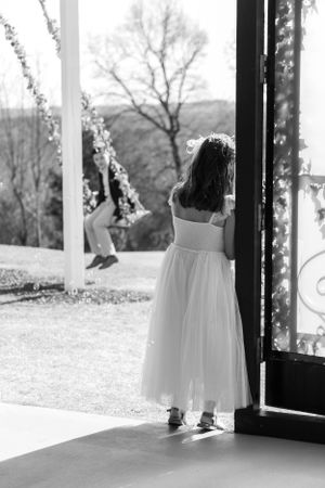 Young flower girl in white dress gazes through doorway decorated with twinkle lights at ring bearer playing on a swing.