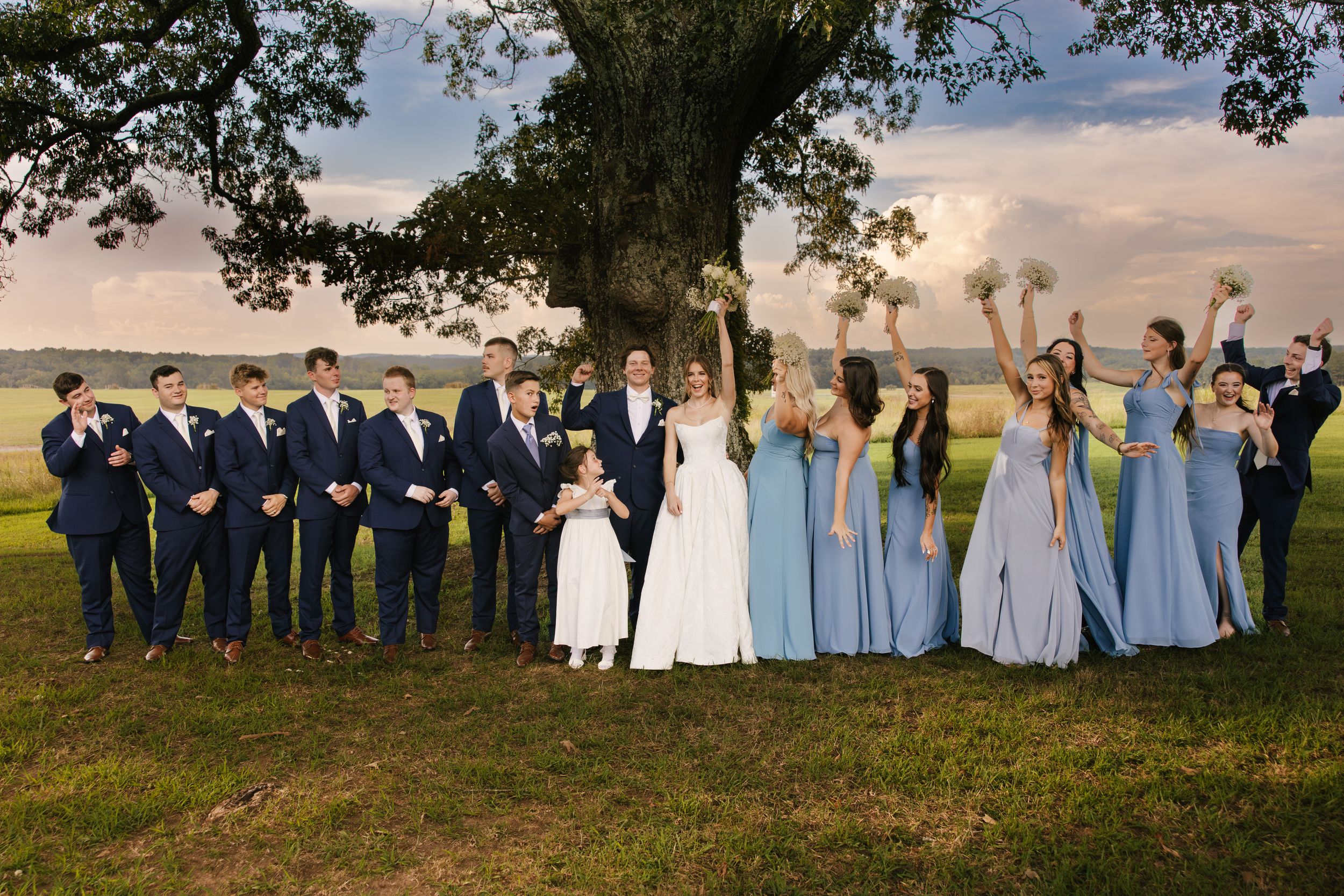 Wedding party poses outdoors under large tree, groomsmen in navy suits and bridesmaids in blue gowns celebrating with bride and groom.