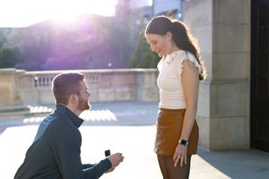 A romantic proposal scene outdoors at sunset with a person kneeling and presenting a ring.