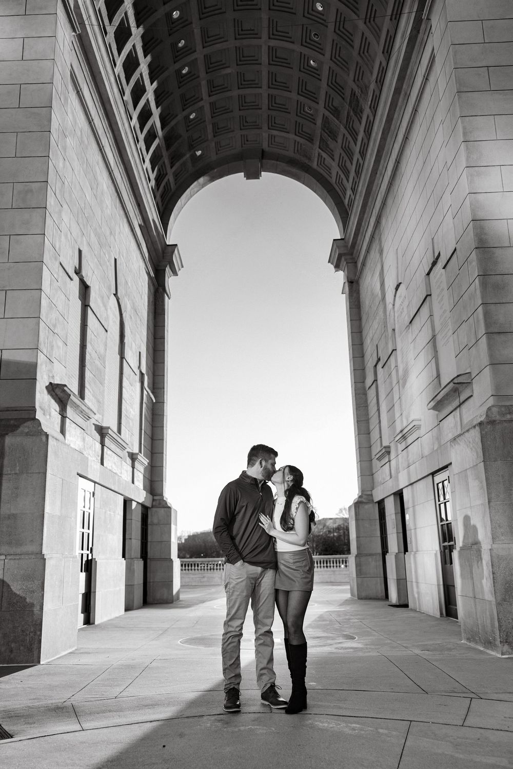 Black and white photo of a couple embracing under a grand stone archway, silhouetted against bright sky.
