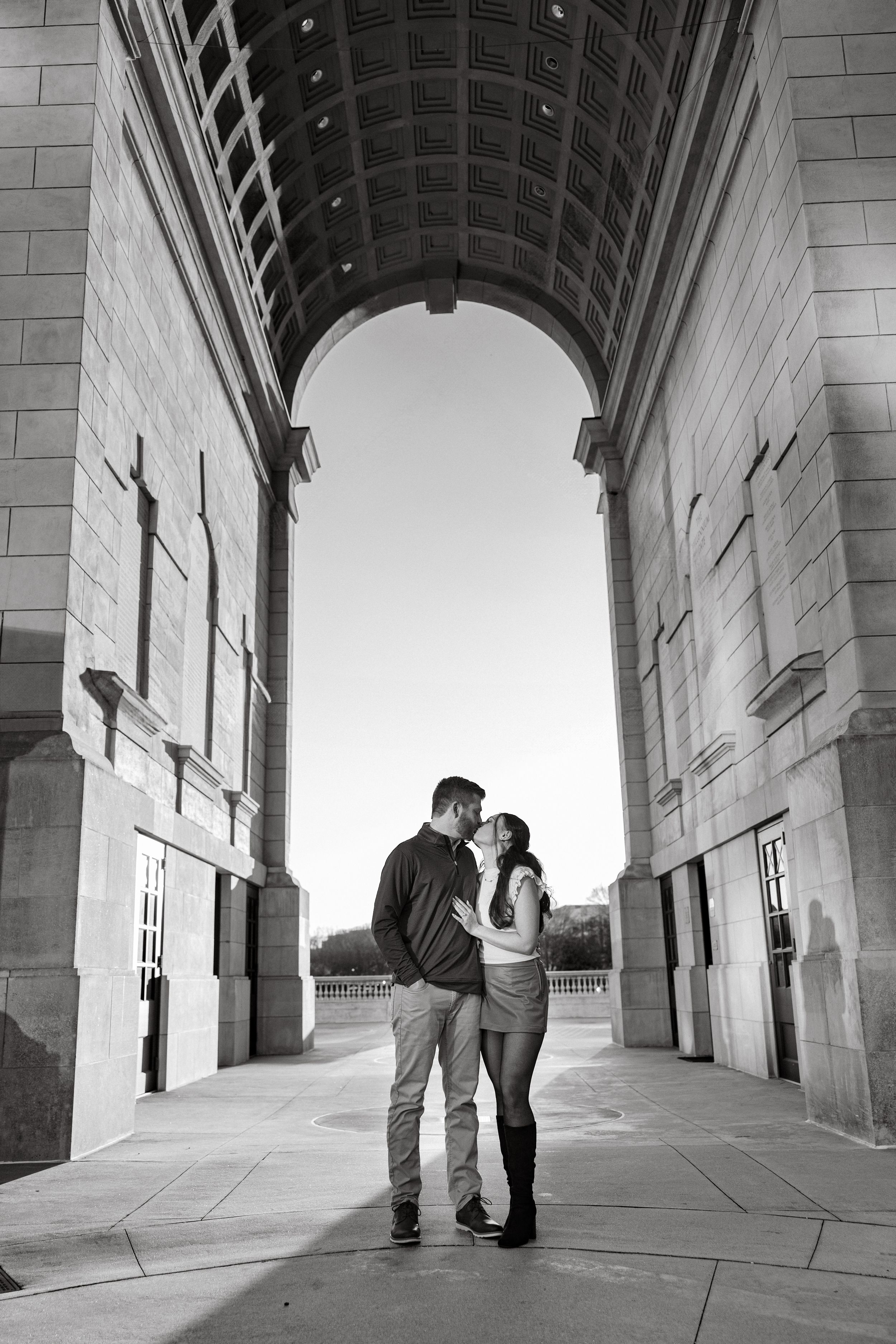 Black and white photo of a couple embracing under a grand stone archway, silhouetted against bright sky.