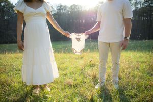 Expecting couple holding baby onesie together in sunny green field, maternity announcement photo outdoors.