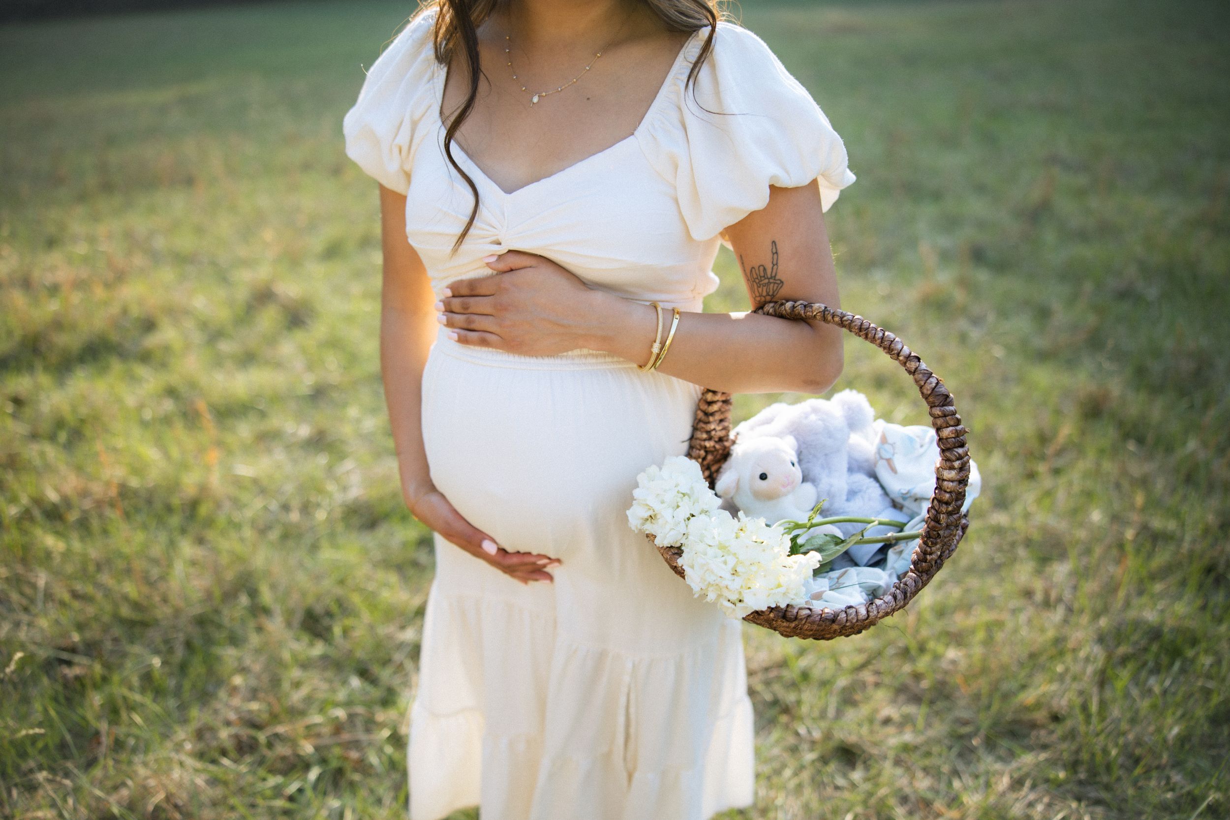 A person in a flowing white dress carries a woven basket filled with flowers in a sunlit meadow.
