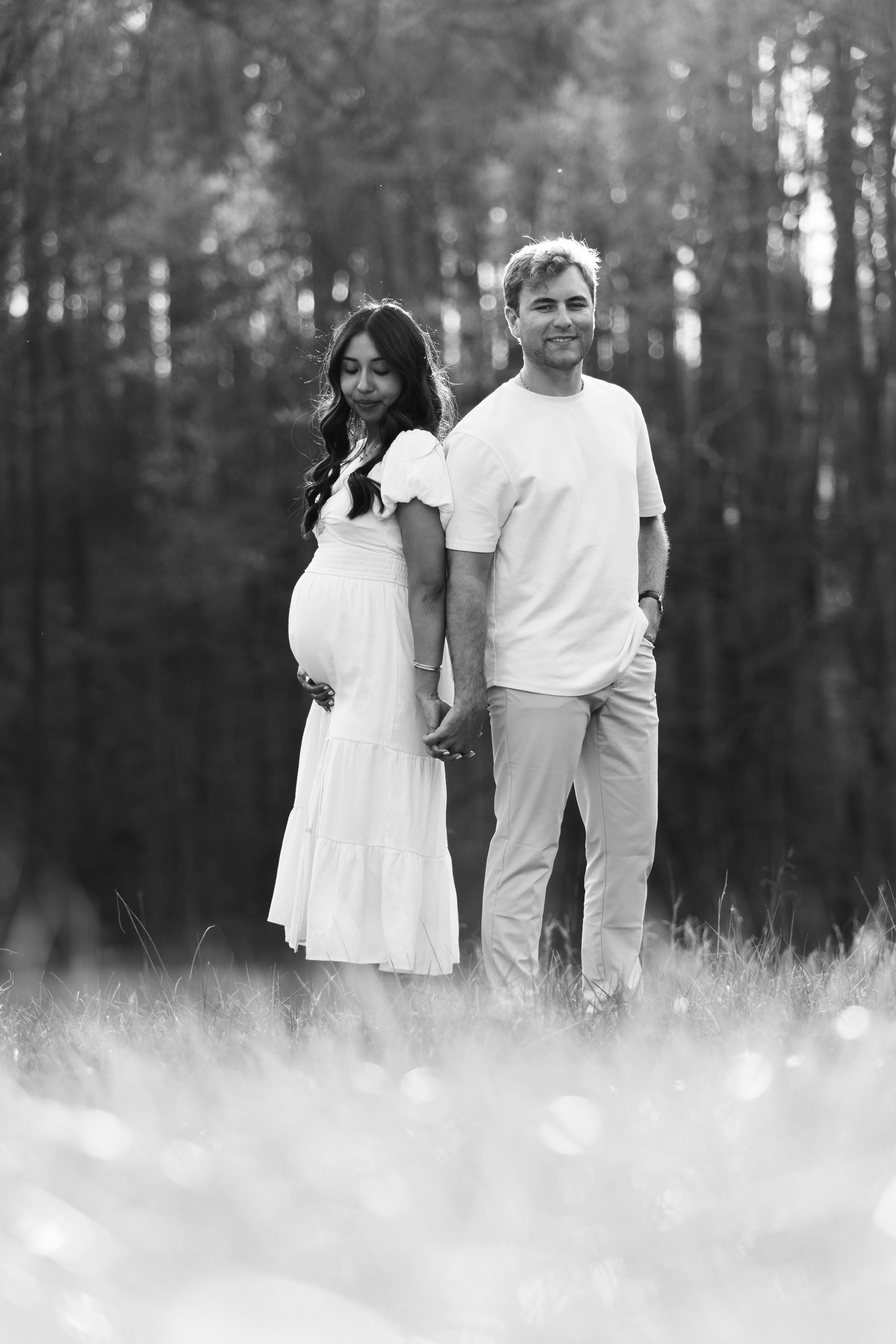 Black and white photo of a pregnant couple holding hands in a field with trees in the background.