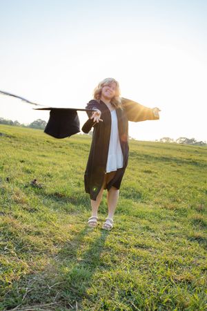 Young girl in graduation gown and cap celebrates outdoors in a sunny green field, arms outstretched with joy.