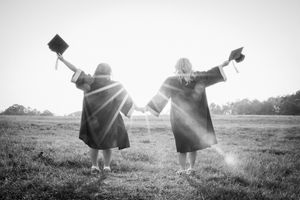 Two graduates in caps and gowns hold hands facing a sunlit field in a black and white photo.