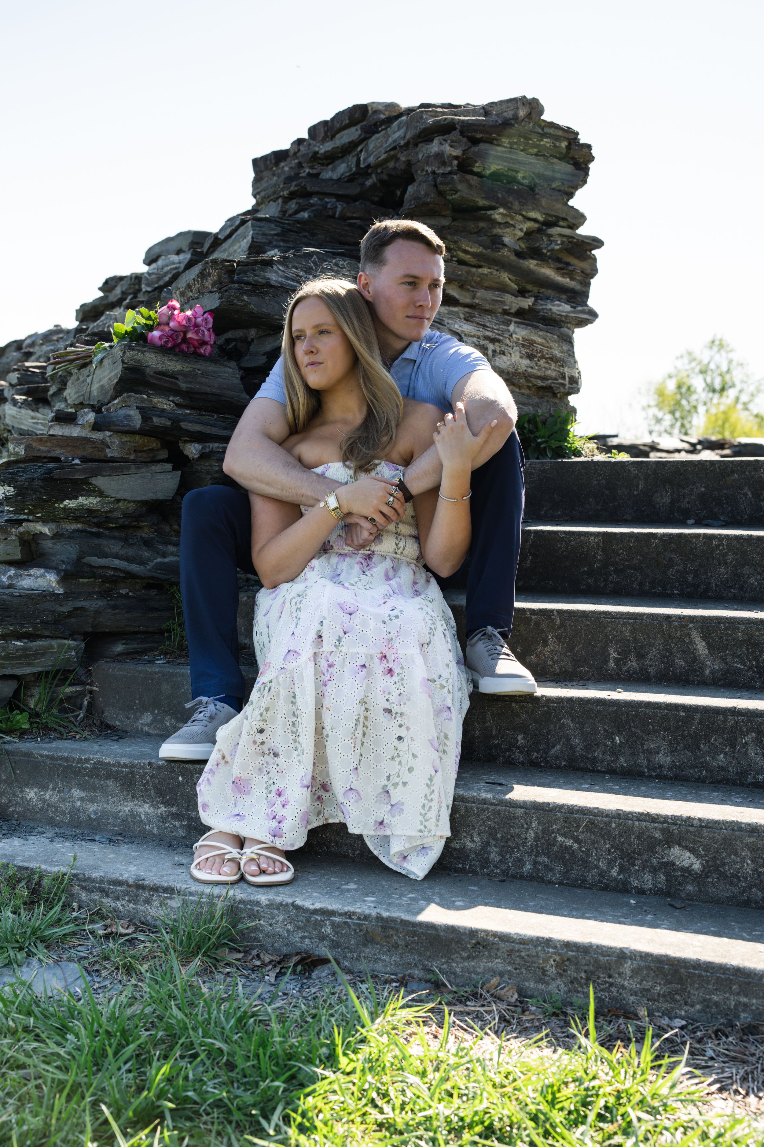 Couple sitting on stone steps, man embracing woman in floral dress, romantic outdoor portrait with rustic rock wall backdrop.