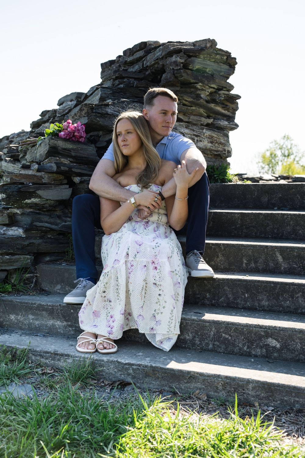 Couple sitting on stone steps, man embracing woman in floral dress, romantic outdoor portrait with rustic rock wall backdrop.