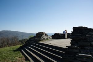 Couple embracing at stone ruins overlook with sweeping mountain valley views under clear blue sky.