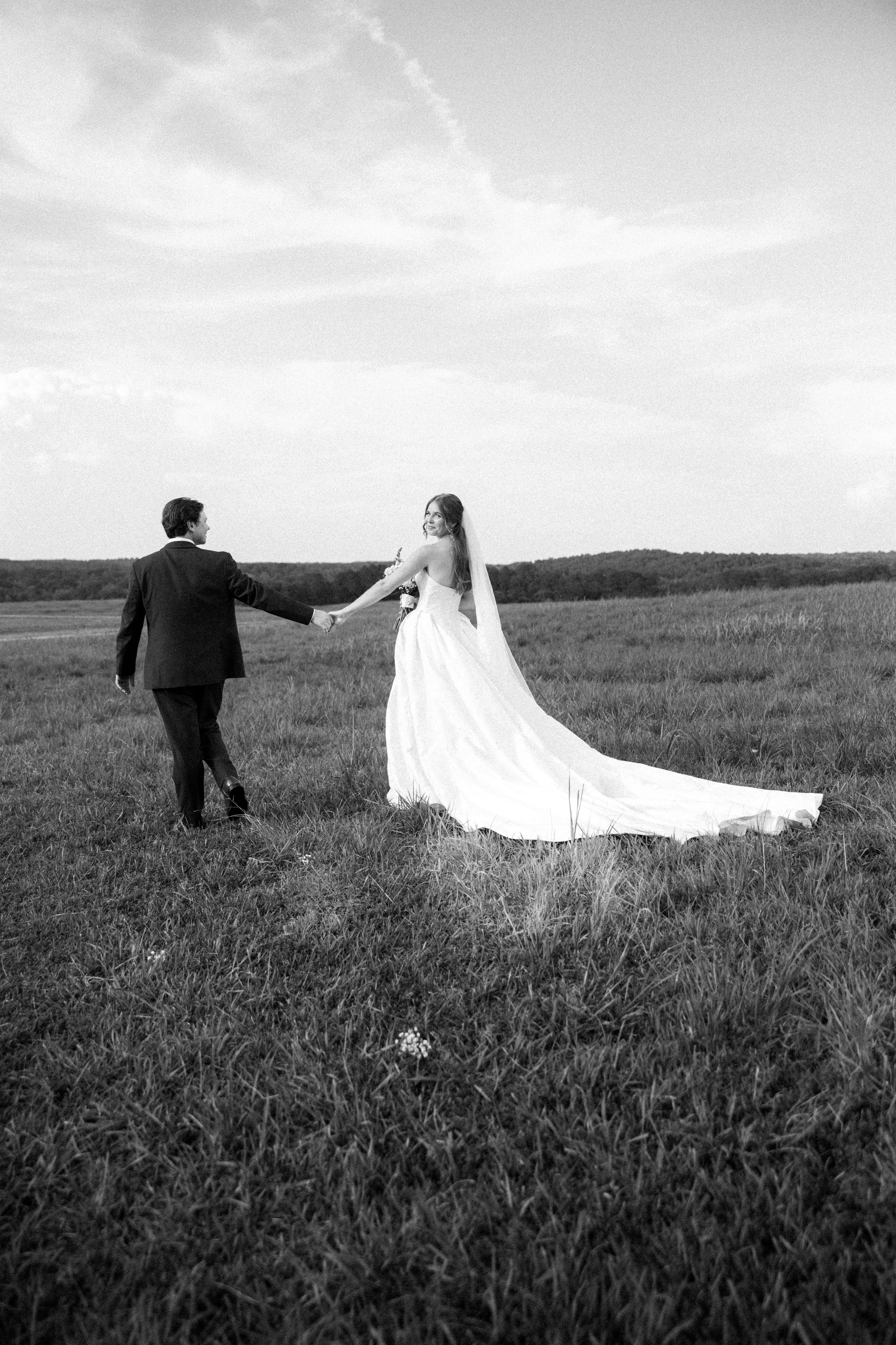 Black and white photo of a bride and groom holding hands walking through a grassy field on their wedding day.
