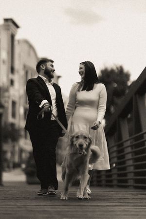 A couple stands on a wooden boardwalk with their fluffy dog in a romantic black and white photo series.